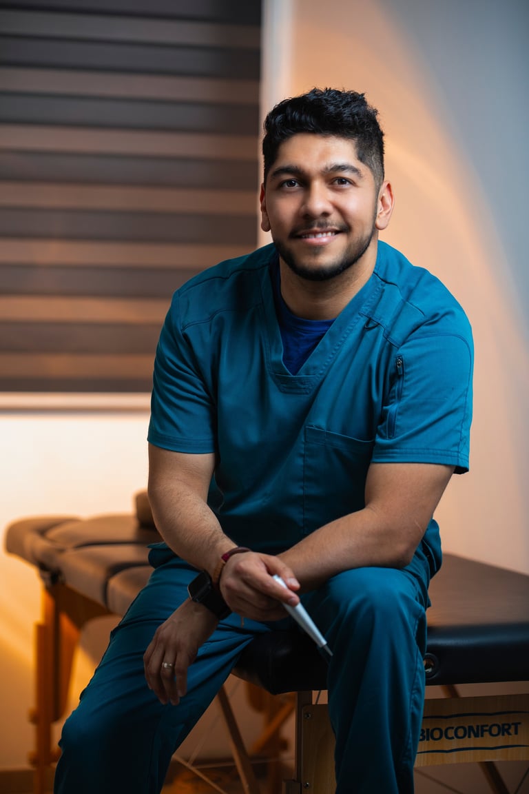 A smiling young man in blue medical scrubs sits on a stool against a modern wall with warm lighting
