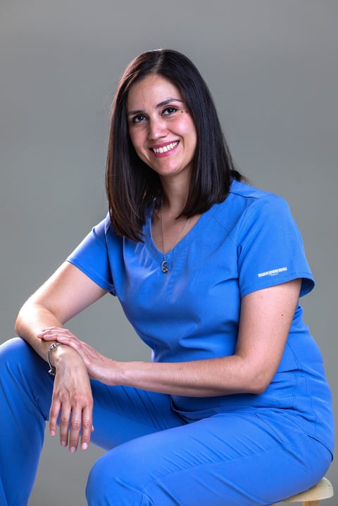 Woman in blue medical scrubs smiling at camera in professional headshot pose against gray background